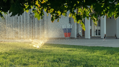 A sprinkler is watering the vibrant green grass under a canopy of trees, with sunlight glistening through the water droplets. In the background, a person rides a scooter near a building with numerous vertical columns, and a sculpture featuring elements of red, white, and blue stands prominently.