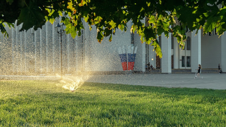 A sprinkler is watering the vibrant green grass under a canopy of trees, with sunlight glistening through the water droplets. In the background, a person rides a scooter near a building with numerous vertical columns, and a sculpture featuring elements of red, white, and blue stands prominently.