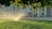 A sprinkler is watering the vibrant green grass under a canopy of trees, with sunlight glistening through the water droplets. In the background, a person rides a scooter near a building with numerous vertical columns, and a sculpture featuring elements of red, white, and blue stands prominently.