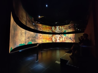 A dimly lit room features a large, curved digital screen displaying ancient or historical symbols and patterns in earthy tones. Several people are seated to the side, observing the display.