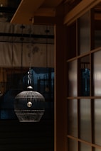 A dimly lit interior with a hanging birdcage-like lamp in the foreground. The warm wooden architecture creates a harmonious contrast with the cool dark tones in the background. Subtle patterns can be seen on the curtains, adding texture to the scene.