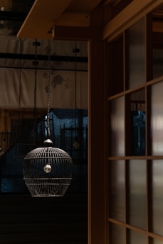 A dimly lit interior with a hanging birdcage-like lamp in the foreground. The warm wooden architecture creates a harmonious contrast with the cool dark tones in the background. Subtle patterns can be seen on the curtains, adding texture to the scene.