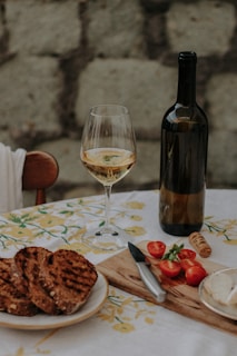 A table is set with a glass of white wine, a bottle, sliced tomatoes, grilled bread, and a cheese spread. The setting has a rustic feel with a stone wall in the background and a white tablecloth with yellow floral patterns.