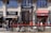 A street scene with multiple storefronts, including a café, a shop, and a currency exchange office. Several pedestrians walk on the sidewalk in front of the buildings. Bicycles are parked in the foreground, and the area is bathed in sunlight.