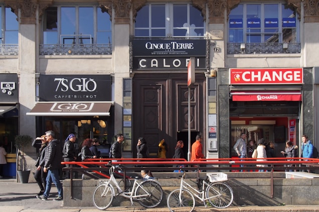 A street scene with multiple storefronts, including a caf&eacute;, a shop, and a currency exchange office. Several pedestrians walk on the sidewalk in front of the buildings. Bicycles are parked in the foreground, and the area is bathed in sunlight.