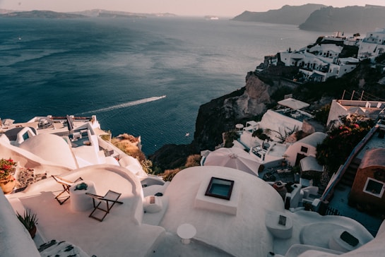 A scenic view of whitewashed buildings perched atop a cliff overlooking a vast expanse of calm blue sea. The architecture features domed roofs and terraces with lounge chairs. Bright flowers add a vibrant contrast to the predominantly white structures. The sea extends to the horizon with a boat creating a trail on the water, while distant islands are visible.