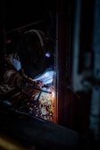 A person equipped with a welding helmet and gloves is engaged in welding, with sparks flying against a dark background.