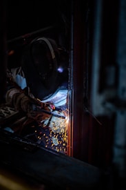 A person equipped with a welding helmet and gloves is engaged in welding, with sparks flying against a dark background.