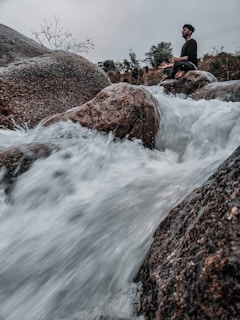 A person is sitting cross-legged in a meditative pose on large rocks beside a fast-flowing stream or waterfall. Surrounding them are trees and a cloudy sky, creating a calm and serene atmosphere amidst the powerful flow of water.