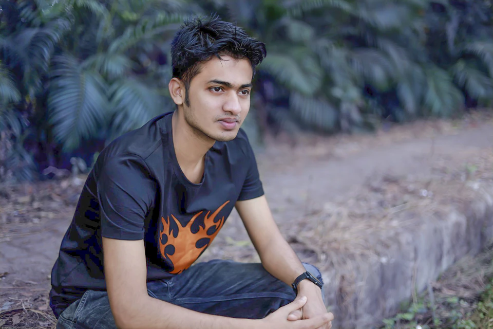 The image shows a young man sitting on a concrete ledge, wearing a black t-shirt with an orange flame design and blue jeans. The man is looking directly at the camera with a serious expression. In the background, there are trees and bushes, creating a natural setting for the scene.