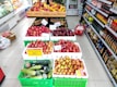A supermarket aisle displays various types of apples and cucumbers in crates. There are red, yellow, and green apples with price tags and a sign indicating a discount. The shelves on the right are stocked with assorted packaged goods and bottles. A chilled section on the left contains more fresh produce.