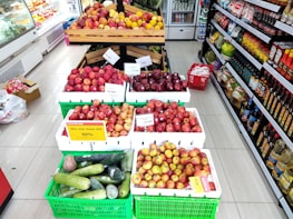 A supermarket aisle displays various types of apples and cucumbers in crates. There are red, yellow, and green apples with price tags and a sign indicating a discount. The shelves on the right are stocked with assorted packaged goods and bottles. A chilled section on the left contains more fresh produce.