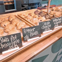 Various types of pastries are displayed on a wooden counter, including croissants and pain au chocolat. The pastries are organized in wooden trays with handwritten chalkboard signs indicating their names and prices. The background features an industrial kitchen setting.