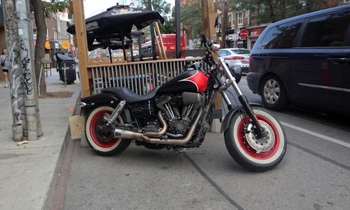 A motorcycle with red and black detailing is parked on the street beside a curb. The background features a wooden outdoor seating area with black umbrellas branded with Sapporo logos. Several vehicles, including a dark minivan and a red bus, are visible on the road. Urban elements such as a trash can, a utility pole covered with stickers, and pedestrians add to the city scene.