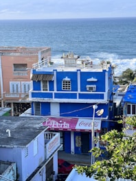 A vibrant coastal scene featuring several colorful buildings beside the ocean. A prominent blue building with white trim and a Coca Cola sign stands in the foreground. To the left, a peach-colored building with white shutters is visible. The ocean waves crash gently in the background, and some greenery can be seen in the foreground.