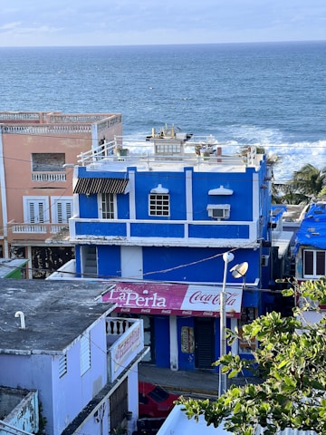 A vibrant coastal scene featuring several colorful buildings beside the ocean. A prominent blue building with white trim and a Coca Cola sign stands in the foreground. To the left, a peach-colored building with white shutters is visible. The ocean waves crash gently in the background, and some greenery can be seen in the foreground.