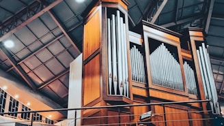 A grand pipe organ with large, polished metal pipes set within a tall wooden structure, situated inside a spacious building with a high ceiling. The architecture features metal beams and a row of windows casting warm light.