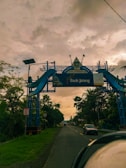 An ornate blue gateway arch spans across a road, with a backdrop of a cloudy sky. The arch features decorative elements and signage including 'KAB. PURWOREJO' and 'Bank Jateng'. Tall trees line the road on both sides, and vehicles are visible driving towards the horizon.