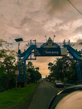 An ornate blue gateway arch spans across a road, with a backdrop of a cloudy sky. The arch features decorative elements and signage including 'KAB. PURWOREJO' and 'Bank Jateng'. Tall trees line the road on both sides, and vehicles are visible driving towards the horizon.