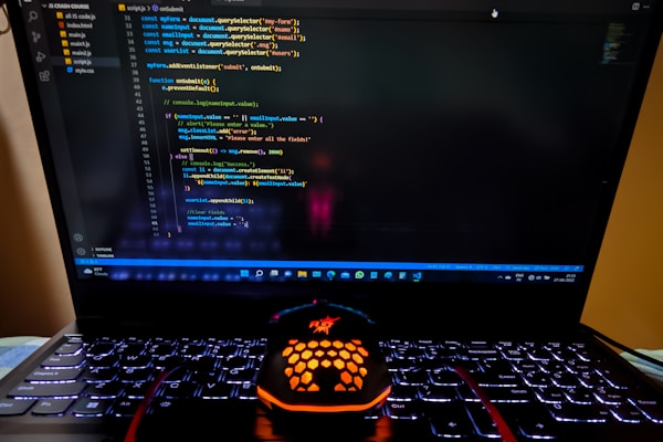 A laptop displaying a coding interface with lines of JavaScript code on the screen. Below the laptop, there is a black and orange gaming mouse with a honeycomb pattern, and its logo is illuminated. The laptop keyboard is backlit, emitting a soft purple glow.