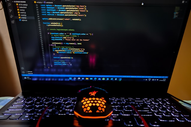 A laptop displaying a coding interface with lines of JavaScript code on the screen. Below the laptop, there is a black and orange gaming mouse with a honeycomb pattern, and its logo is illuminated. The laptop keyboard is backlit, emitting a soft purple glow.