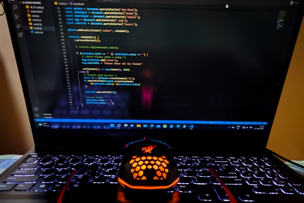 A laptop displaying a coding interface with lines of JavaScript code on the screen. Below the laptop, there is a black and orange gaming mouse with a honeycomb pattern, and its logo is illuminated. The laptop keyboard is backlit, emitting a soft purple glow.