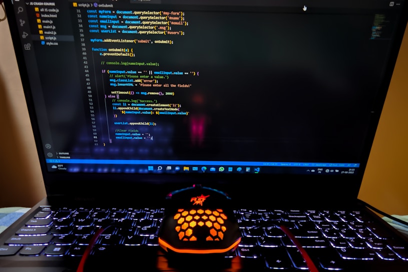 A laptop displaying a coding interface with lines of JavaScript code on the screen. Below the laptop, there is a black and orange gaming mouse with a honeycomb pattern, and its logo is illuminated. The laptop keyboard is backlit, emitting a soft purple glow.