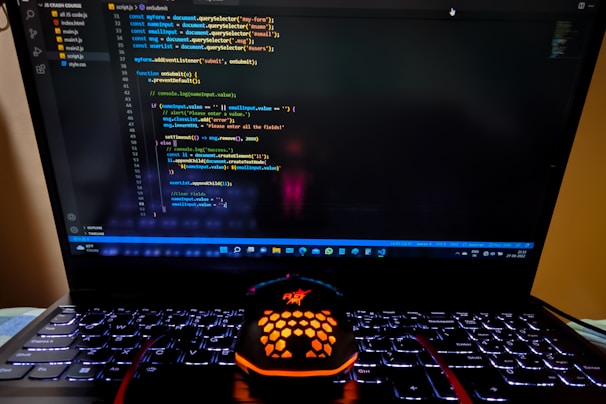 A laptop displaying a coding interface with lines of JavaScript code on the screen. Below the laptop, there is a black and orange gaming mouse with a honeycomb pattern, and its logo is illuminated. The laptop keyboard is backlit, emitting a soft purple glow.