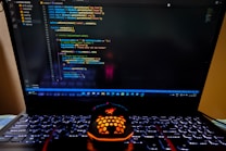 A laptop displaying a coding interface with lines of JavaScript code on the screen. Below the laptop, there is a black and orange gaming mouse with a honeycomb pattern, and its logo is illuminated. The laptop keyboard is backlit, emitting a soft purple glow.