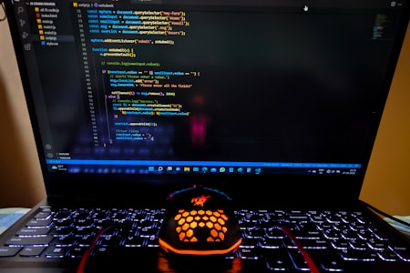 A laptop displaying a coding interface with lines of JavaScript code on the screen. Below the laptop, there is a black and orange gaming mouse with a honeycomb pattern, and its logo is illuminated. The laptop keyboard is backlit, emitting a soft purple glow.