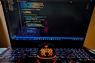A laptop displaying a coding interface with lines of JavaScript code on the screen. Below the laptop, there is a black and orange gaming mouse with a honeycomb pattern, and its logo is illuminated. The laptop keyboard is backlit, emitting a soft purple glow.
