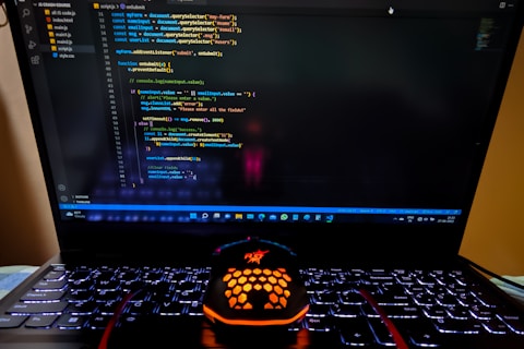 A laptop displaying a coding interface with lines of JavaScript code on the screen. Below the laptop, there is a black and orange gaming mouse with a honeycomb pattern, and its logo is illuminated. The laptop keyboard is backlit, emitting a soft purple glow.