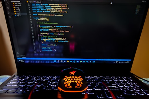 A laptop displaying a coding interface with lines of JavaScript code on the screen. Below the laptop, there is a black and orange gaming mouse with a honeycomb pattern, and its logo is illuminated. The laptop keyboard is backlit, emitting a soft purple glow.