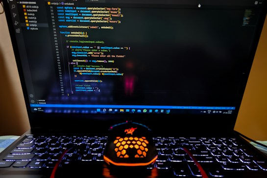 A laptop displaying a coding interface with lines of JavaScript code on the screen. Below the laptop, there is a black and orange gaming mouse with a honeycomb pattern, and its logo is illuminated. The laptop keyboard is backlit, emitting a soft purple glow.