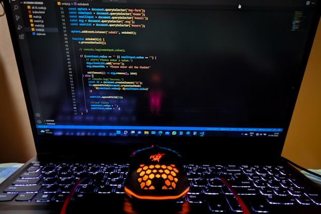 A laptop displaying a coding interface with lines of JavaScript code on the screen. Below the laptop, there is a black and orange gaming mouse with a honeycomb pattern, and its logo is illuminated. The laptop keyboard is backlit, emitting a soft purple glow.