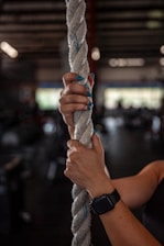 A hand wearing a smartwatch grips a thick rope firmly in a dimly lit gym setting. The background is blurred, indicating gym equipment and large windows that let in natural light. The nails are painted teal, matching the few strands of blue in the rope.