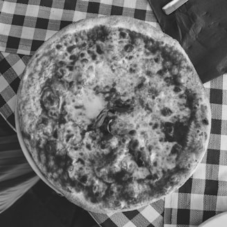 A pizza with a crisp crust and melted cheese is placed on a checkered tablecloth. There is a sprig of basil in the center of the pizza. A napkin and a piece of silverware are partially visible on the table.