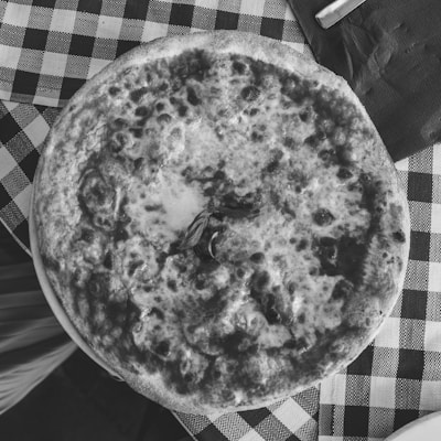 A pizza with a crisp crust and melted cheese is placed on a checkered tablecloth. There is a sprig of basil in the center of the pizza. A napkin and a piece of silverware are partially visible on the table.