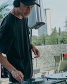 A person wearing a black t-shirt and black cap is standing outdoors, using DJ equipment. The surroundings appear to be a terrace or outdoor space with some greenery and buildings in the background.