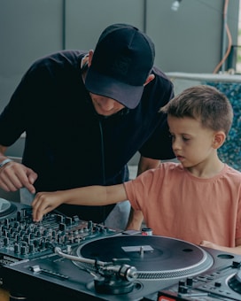 An adult and a child are interacting with DJ equipment. The adult is wearing a black cap and shirt while assisting the child, who is wearing a pink shirt, in operating the turntables.