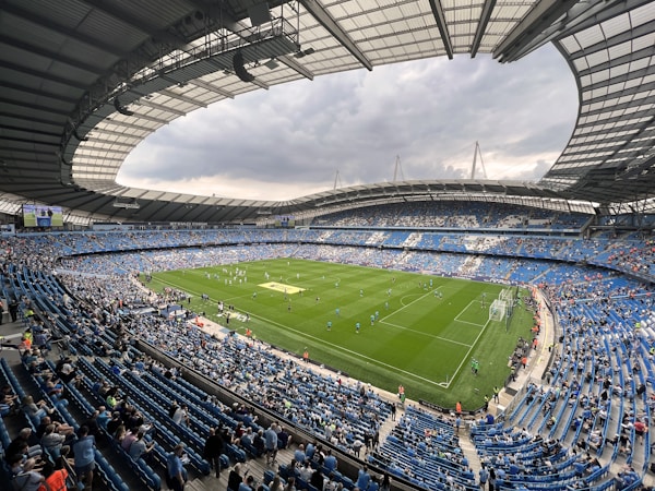 A large football stadium filled with spectators, featuring a green pitch with players preparing for a match. The stadium is covered with a partially transparent roof, allowing natural light. The seating is predominantly blue, and the weather appears cloudy, casting a subdued light over the field.
