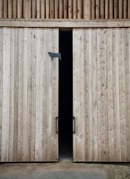 Two tall wooden barn doors with a small gap between them, revealing a dark interior. The doors are made of vertical wooden planks with visible grain patterns and have metal handles. A small silhouette of a cow is attached to one door as a decoration.