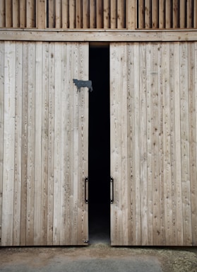 Two tall wooden barn doors with a small gap between them, revealing a dark interior. The doors are made of vertical wooden planks with visible grain patterns and have metal handles. A small silhouette of a cow is attached to one door as a decoration.
