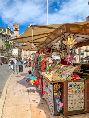 A lively market stall displaying a variety of colorful souvenirs, including Venetian masks, keychains, and small toys. The stall is set against a picturesque backdrop of historic European architecture. People are walking nearby, enjoying the bustling street atmosphere under a blue sky with some clouds.