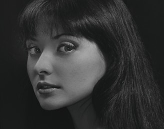 A black and white portrait of a woman with long dark hair looking over her shoulder. Her expression appears thoughtful and serene, with eyes gazing directly at the viewer. The lighting highlights her facial features against a dark background.