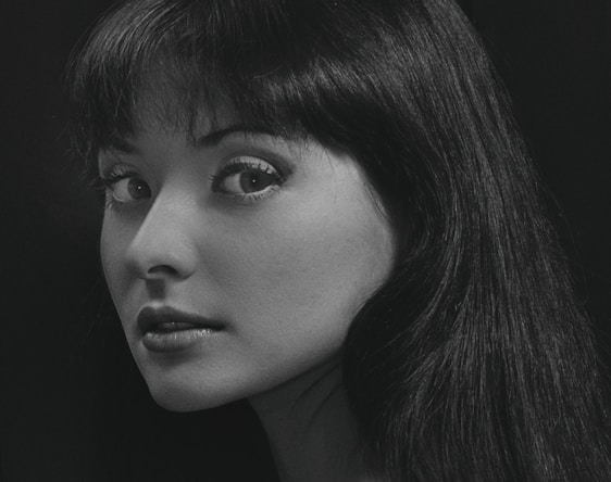 A black and white portrait of a woman with long dark hair looking over her shoulder. Her expression appears thoughtful and serene, with eyes gazing directly at the viewer. The lighting highlights her facial features against a dark background.