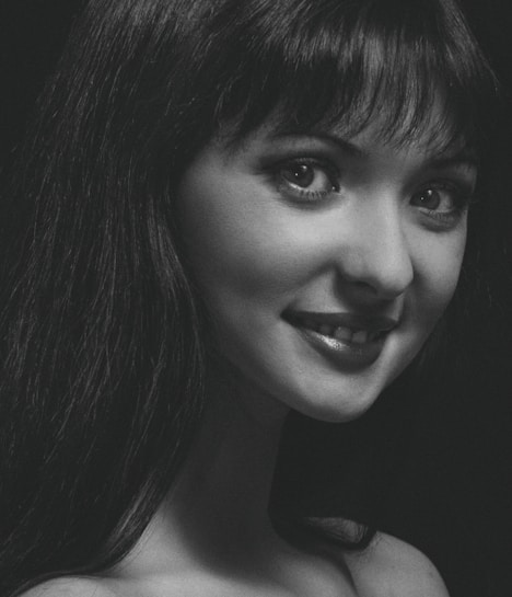 A monochrome portrait of a woman with long dark hair, displaying a soft smile and looking towards the camera. The lighting emphasizes her facial features, creating a dramatic contrast with the dark background.