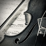 A close-up view of a violin intersecting with a sheet of music. The strings and body of the violin contrast with the musical notes printed on the page. The scene is captured in black and white, highlighting the textures and details of the wood grain and inked sheet.