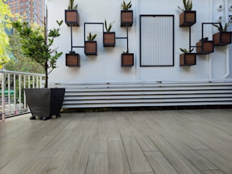 A modern outdoor space features a wall with geometric black metal frames that hold small wooden planters, each containing a plant. There is a grid panel next to them on a white wall. A large black planter with a small tree is placed on a wooden-tiled floor, and a white railing is visible on the left side, with trees and a building in the background.