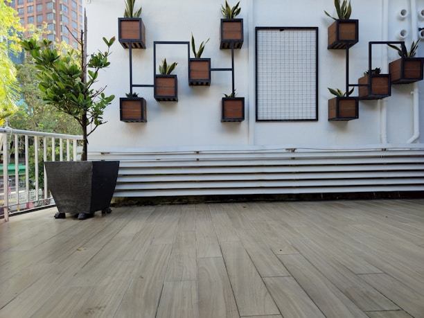 A modern outdoor space features a wall with geometric black metal frames that hold small wooden planters, each containing a plant. There is a grid panel next to them on a white wall. A large black planter with a small tree is placed on a wooden-tiled floor, and a white railing is visible on the left side, with trees and a building in the background.